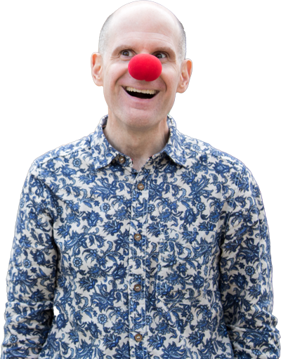 Luke Perone wearing a patterned blue and white shirt smiles broadly, sporting a large red clown nose. The background, blurred with horizontal stripes, hints at an improv setting that adds a touch of social therapeutic charm to the scene.