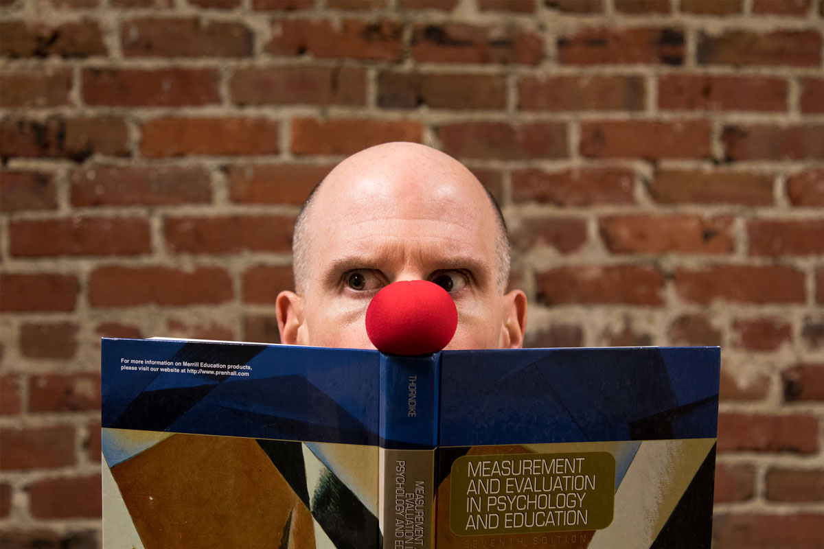 Luke Perone with a red clown nose, reminiscent of an improv performer, is holding and partially hiding behind a book against a brick wall backdrop. The book is titled "Measurement and Evaluation in Psychology and Education." Only his eyes and nose are visible, adding a touch of social therapeutic humor.