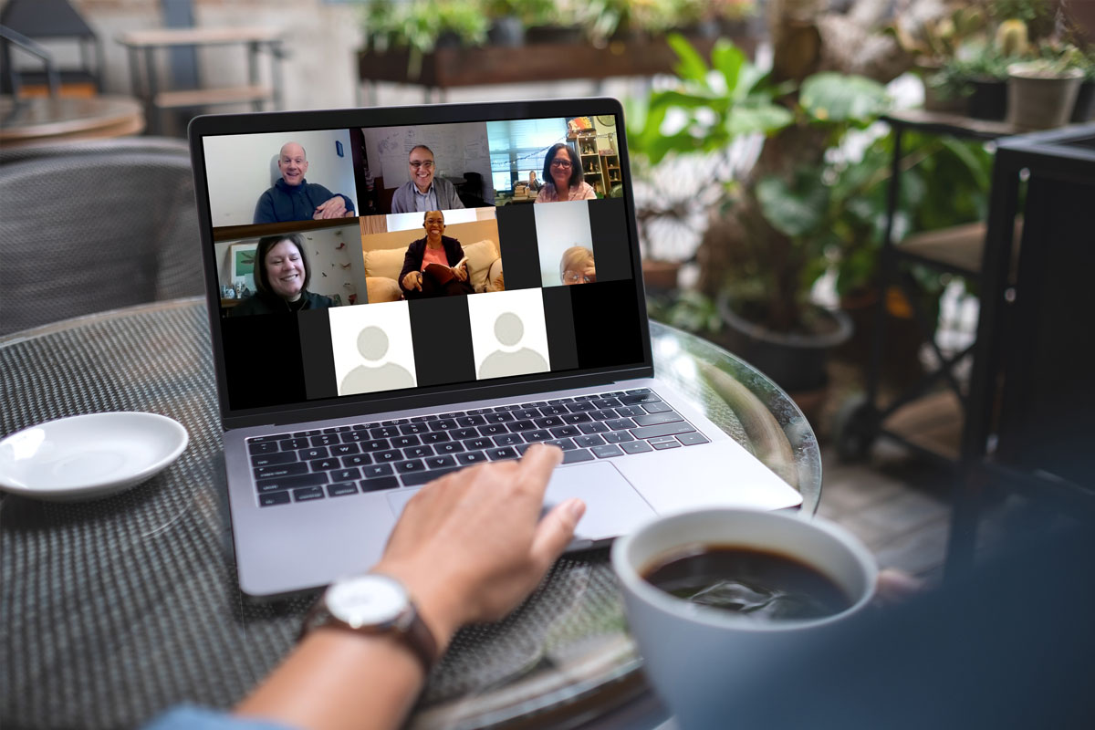 A person holding a cup of coffee participates in a consulting video conference with six others displayed on a laptop screen. Each participant appears in their own video box, and two boxes display user icons instead of faces. The cafe, adorned with plants, offers a cozy setting.