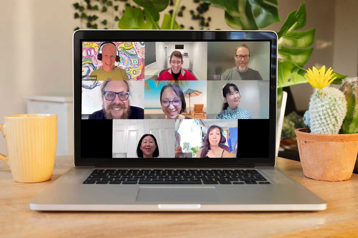 A laptop screen shows a virtual meeting with eight smiling participants in individual windows, engaging in a social therapeutic session. The laptop is on a wooden table with a yellow mug and a small potted cactus. There are green plants in the background.