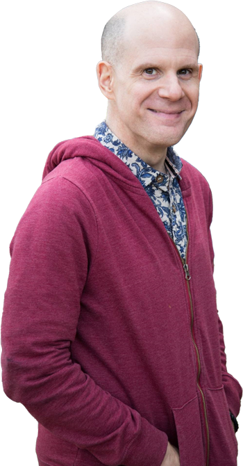 Luke Perone, wearing a patterned shirt under a maroon zip-up hoodie stands smiling with his hands in his pockets. The background, reminiscent of an improv stage, features blurred colors in shades of grey, blue, and brown.