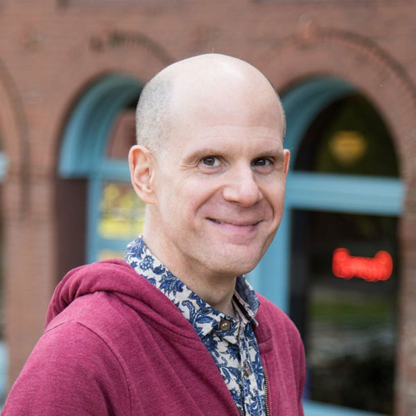 Luke Perone smiles at the camera. He is wearing a red hoodie over a patterned shirt. The background shows a building with arched windows painted blue, and a lighted neon sign is visible, hinting at an improv club known for its social therapeutic impact.