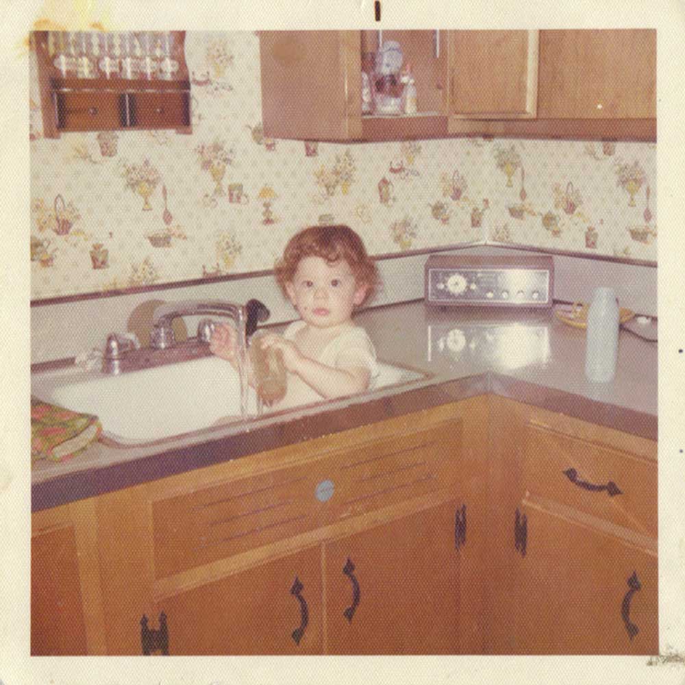 A young child with curly hair is sitting in a kitchen sink, playing with the water from the faucet like an improv actor. The kitchen has wooden cabinets and floral wallpaper. Various kitchen items are on the counter, and an old-fashioned radio provides a social therapeutic ambiance in the background.
