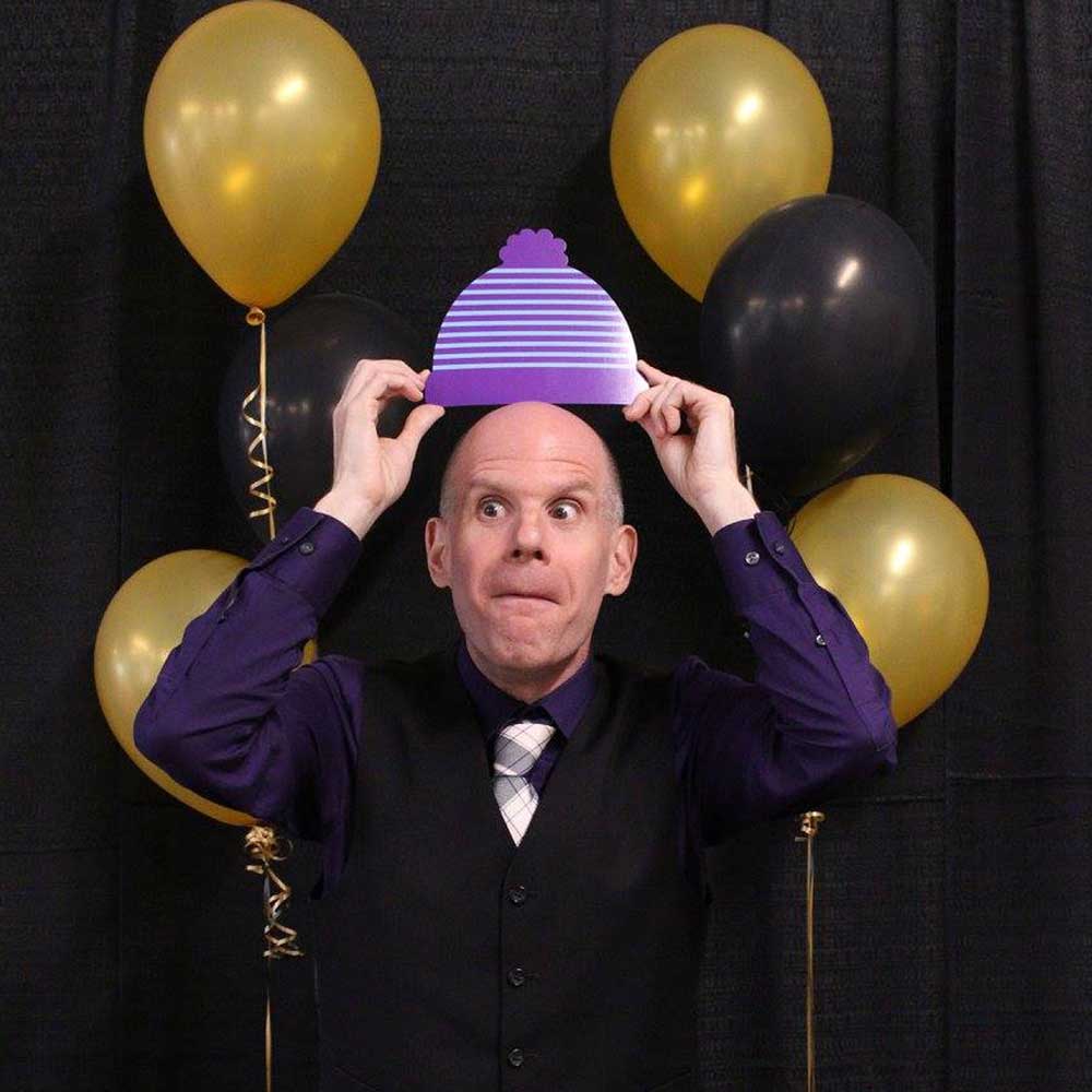 Luke Perone standing in front of black and gold balloons, holding a purple and white striped hat above his head. He is wearing a purple shirt, black vest, and patterned tie, and has a playful expression on his face. His pose suggests an improv performance.