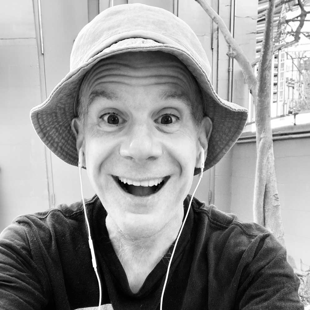 Black and white photo of a cheerful Luke Perone wearing a bucket hat and earphones, smiling widely at the camera. The outdoor setting with trees and a building structure adds to the scene’s charm, evoking a sense of joy, possibly from an improv session.