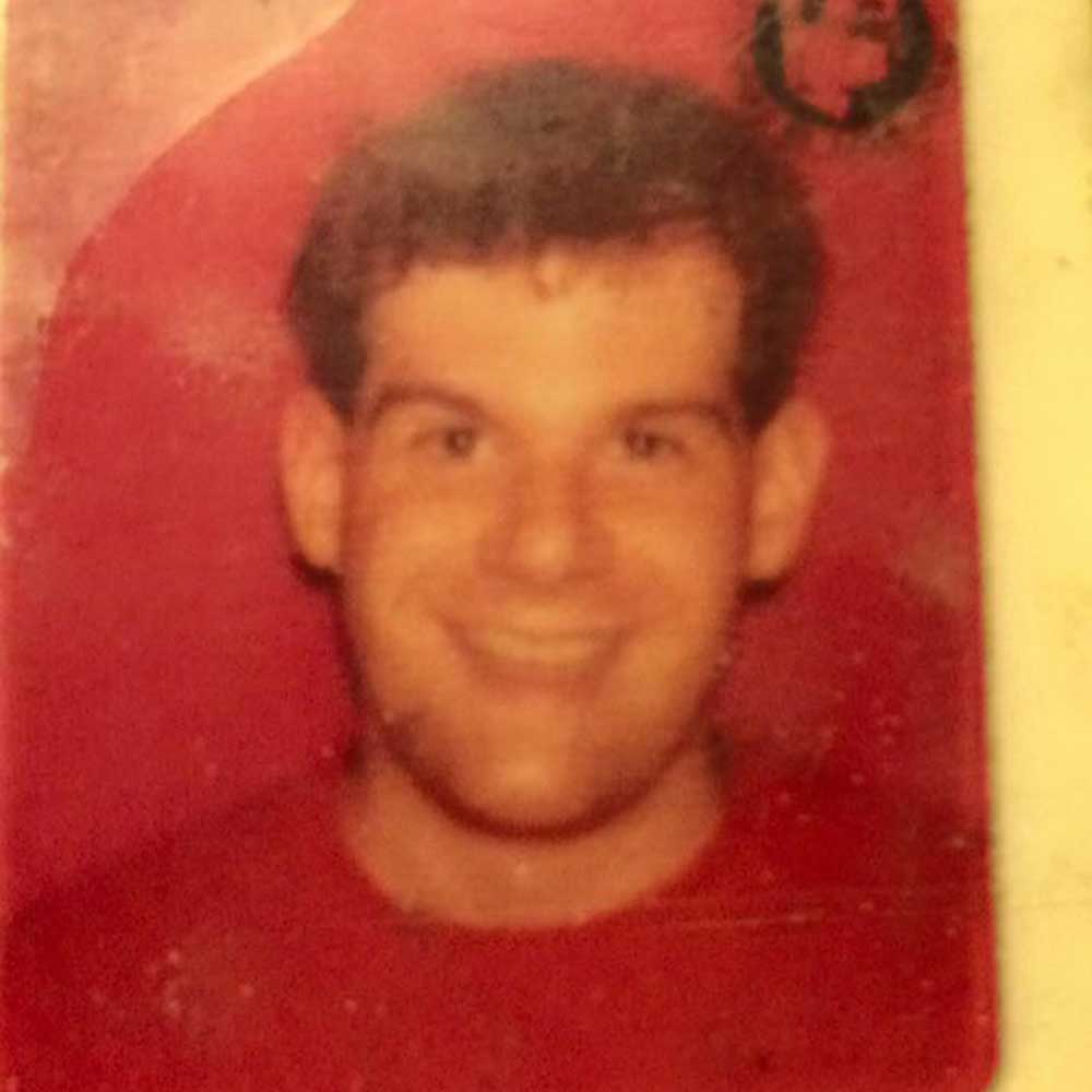 A close-up photo of a young man with short dark hair, smiling broadly at the camera. The background is red, and the image appears to be a slightly worn identification picture, perhaps taken during one of his social therapeutic improv sessions.