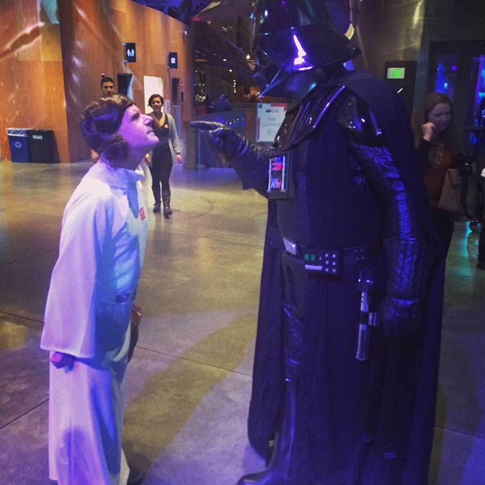 A person dressed as Princess Leia is standing defiantly, leaning forward, while another person dressed as Darth Vader points at them. This social therapeutic improv interaction is taking place in a well-lit, indoor environment with other individuals and decorations visible in the background.
