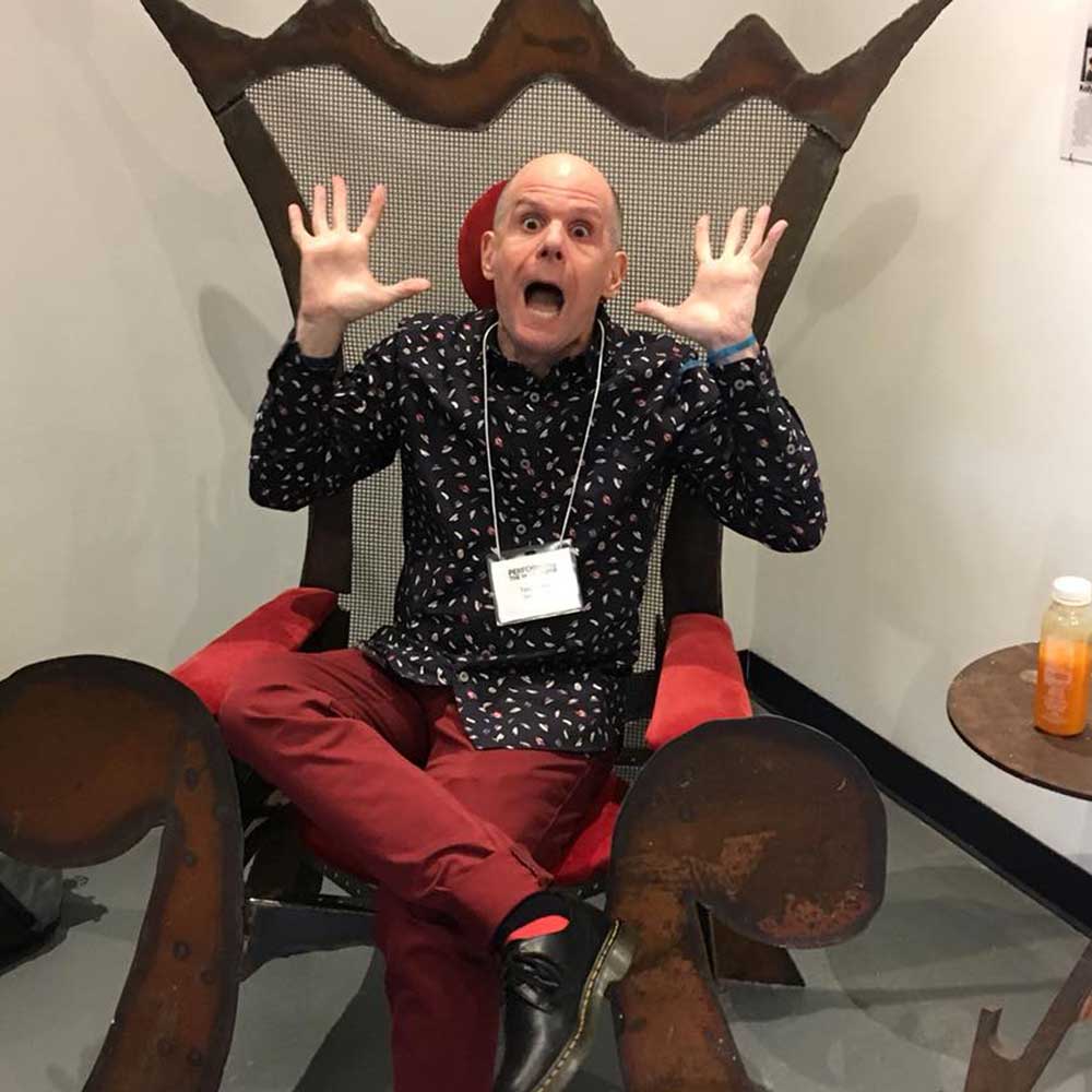 Luke Perone wearing a patterned black shirt and red pants sits on a large, ornate chair with his hands raised in an exaggerated surprised expression, reminiscent of an improv performance. An orange drink bottle is on a small side table to his right.