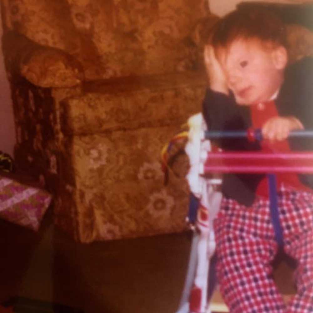 A young child is sitting in a baby walker indoors, covering their face with one hand. Wearing a dark jacket and red plaid pants, the little one seems to be playing an improv game. Behind the child is a floral-patterned armchair, with a wrapped gift on the floor to the left.