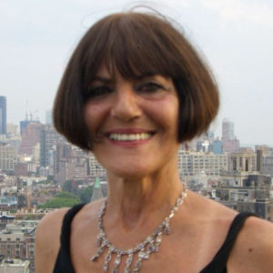 A woman with a short brown bob hairstyle is smiling in an outdoor setting. She is wearing a black top and a silver necklace. The background features a cityscape with numerous buildings under a partly cloudy sky, providing the perfect backdrop for her social therapeutic improv session.