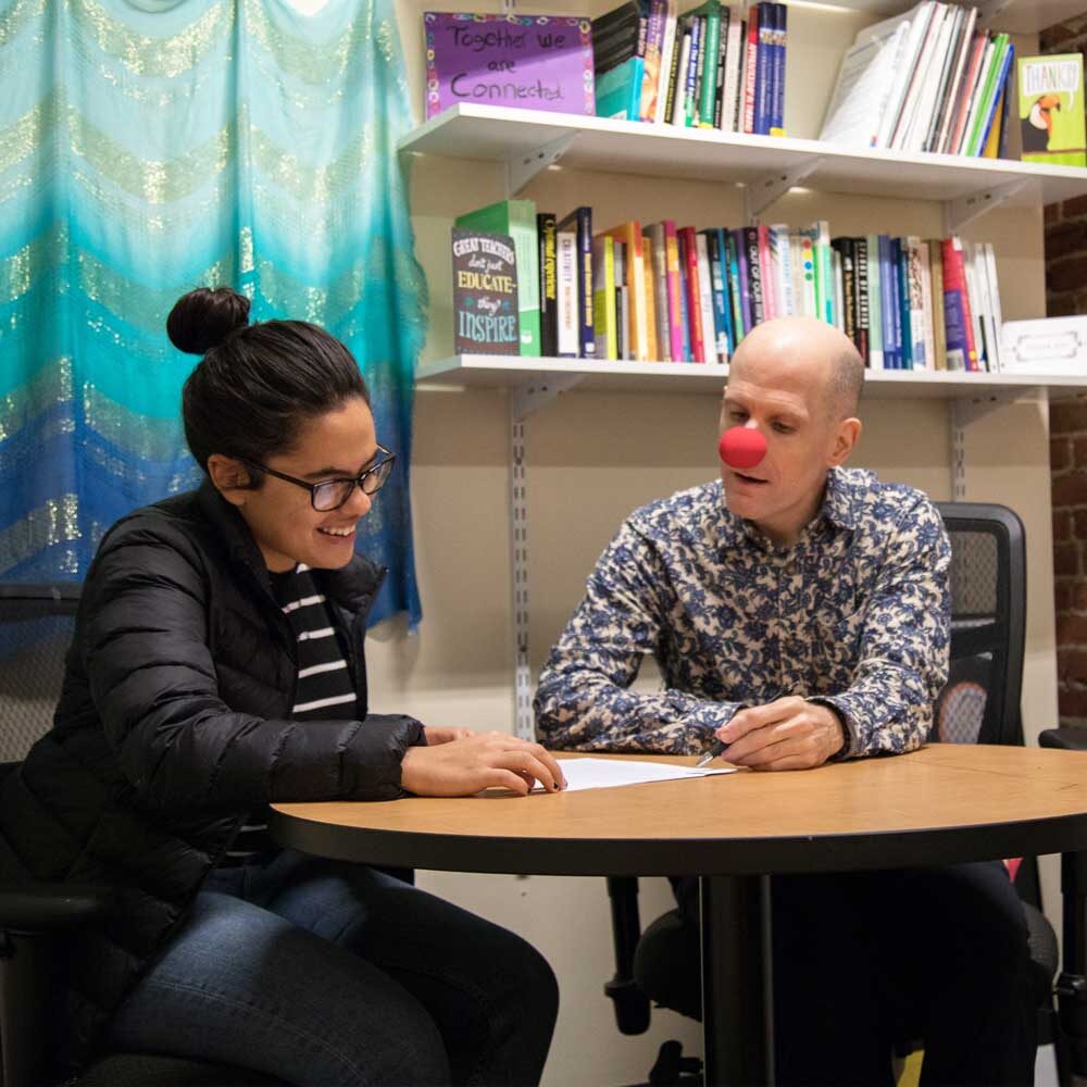 A woman and Luke Perone with a red clown nose, hinting at a coaching session, sit at a round table in an office. The man is writing on a piece of paper while the woman smiles and looks on. Behind them are bookshelves filled with colorful books and a blue curtain, adding a social therapeutic vibe to the scene.